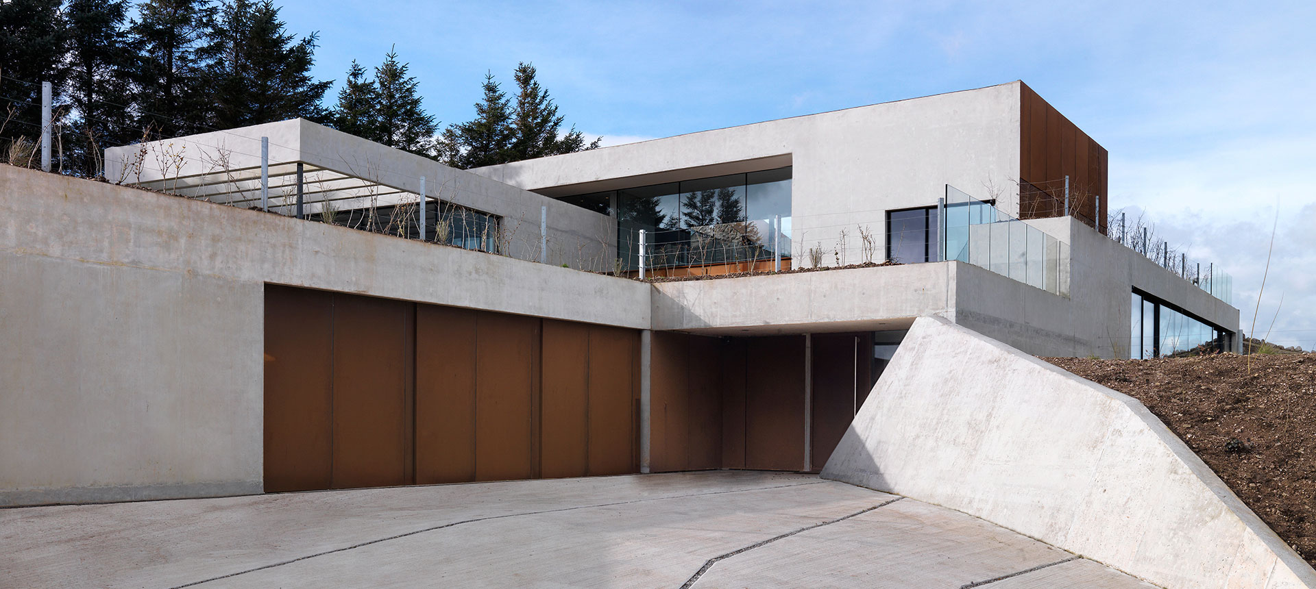 A modern residence with geometric design, combining concrete and wood, featuring large windows and a flat roof, under a partly cloudy sky