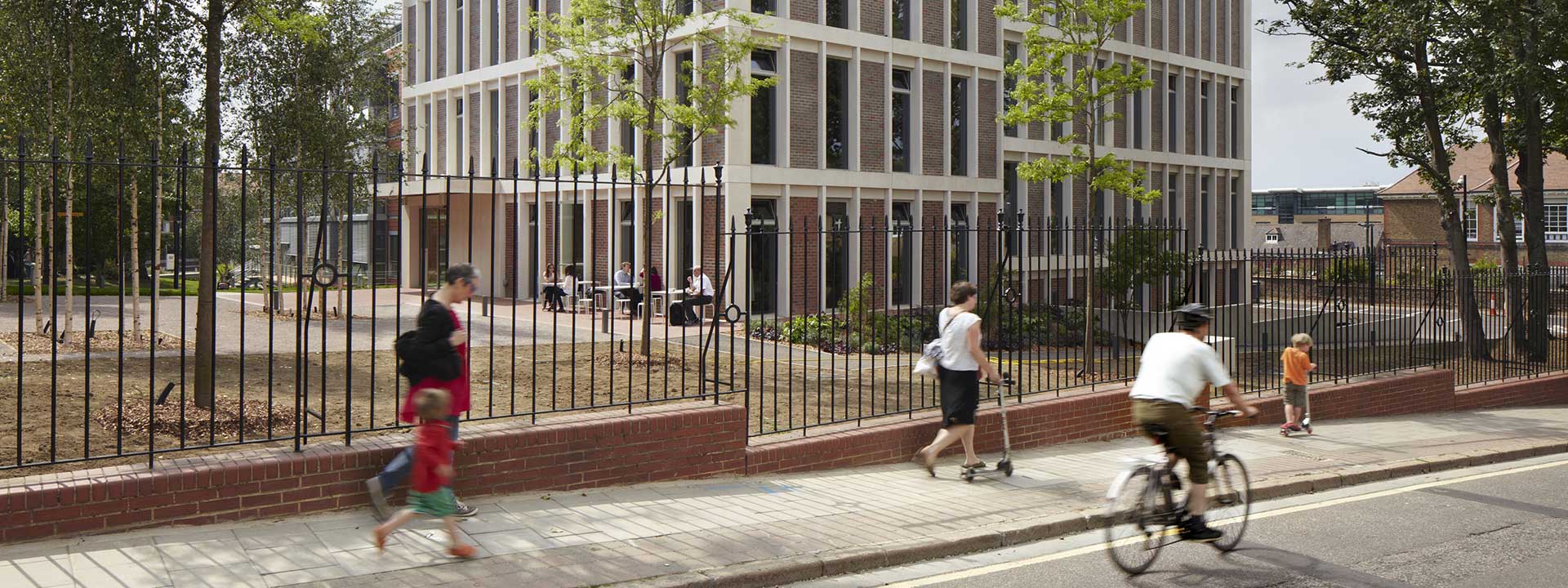 Three-storey building with floor-to-ceiling windows, with people sitting at tables in front of it. Around the house is a courtyard with trees, the house is surrounded by a fence, in front of it people are walking on the pavement and the street. 